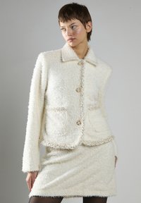 Hvid, struktureret jakke med krave, forlommer og dekorative knapper. Kombineret med en matchende struktureret nederdel, begge fremstillet i et blødt, fluffy materiale.