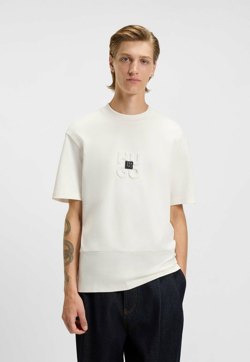 Kortærmet hvid t-shirt lavet af glat stof, med et forhøjet "HUGO" logo i midten og en afslappet pasform. Båret med mørke denim bukser.