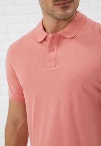 Polo majica od mekanog koraljnog materijala s teksturiranim završetak, s ovratnikom i placketom s tri gumba, dizajnirana za prilagođeni izgled.
