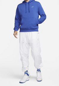 Felpa blu in pile con tasca frontale, pantaloni della tuta bianchi larghi e sneakers bianche con dettagli blu e polsini a coste. Logo Nike sul petto.
