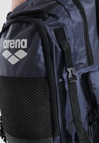 Mørkeblå rygsæk med netpanel, sorte accenter og flere lynlåse. Funktioner inkluderer logo og justerbare stropper til bæring. Holdbart materiale.