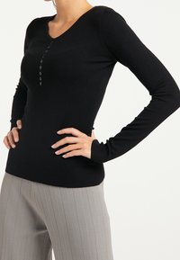 Maglietta nera a maniche lunghe in tessuto morbido con scollatura a V e una fila di bottoni neri sul davanti, abbinata a pantaloni gray a righe sottili.