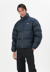 Morska puffer jakna s visokim ovratom, prednjim zatvaranjem na zip i elastičnim manžetama. Ima mali logo na grudima i kviltanu teksturu.