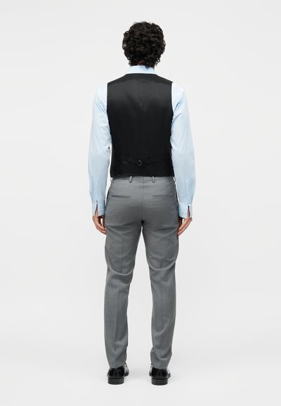Άνδρας με σγουρά μαλλιά, φοράει μαύρο γιλέκο πάνω από ανοιχτό μπλε πουκάμισο και γκρι επίσημο παντελόνι, στέκεται γυρισμένος με την πλάτη σε λευκό απλό φόντο.