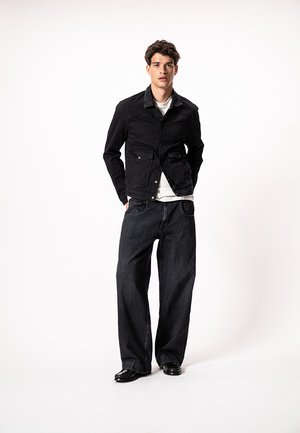 Νέος άνδρας που στέκεται, φοράει μαύρο μπουφάν, λευκό πουκάμισο, φαρδιά μαύρα τζιν και μαύρα παπούτσια, σε λευκό φόντο.