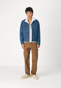 Chaqueta de mezclilla con un patrón azul, camisa blanca con cuello, pantalones rectos marrones y zapatillas de crema. Botones visibles en la chaqueta.