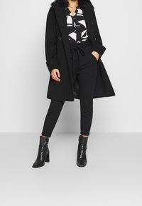 Svart dubbelsidig kappa med krage i konstpäls, svart mönstrad blus, svarta byxor och stövlar med dragkedja vid ankeln. Platt skosilhuett.