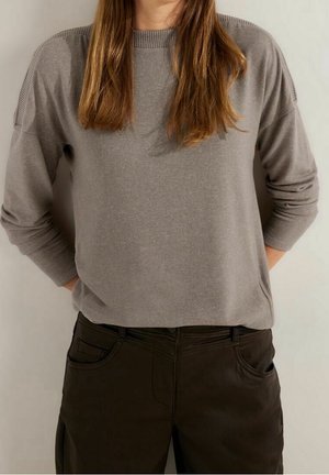 Vrouw draagt een lichtbruine trui met lange mouwen en geribbelde boorden en donkere bruine broek, staat met haar handen achter haar rug.