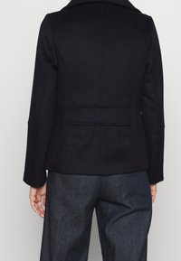 Chaqueta de mezcla de lana en un oscuro azul marino con un corte ajustado, hombros estructurados y una textura suave. Presenta una abertura en la parte posterior y mangas largas.