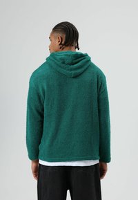 Sweat à capuche vert et duveteux avec une surface texturée ; manches longues ; coupe décontractée ; porté par-dessus une chemise blanche, vu de dos.