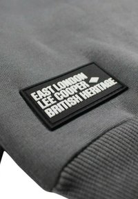 Sweat-shirt gris en tissu texturé. Présente une étiquette noire rectangulaire avec un texte blanc indiquant "EAST LONDON LEE COOPER BRITISH HERITAGE."