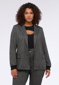 Šedý pruhovaný blazer se dvěma předními kapsami, zapínáním na knoflík a černými žebrovanými manžetami, nošený přes černou košili s výřezem.