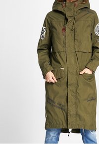 Olivgrön lång jacka med huva, med fickor och broderad text. Tillverkad av hållbart material med rak skärning och justerbara element.