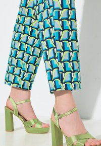 Piernas con pantalones cortos con patrones geométricos en azul, verde y blanco, y sandalias verdes de tacón alto con tiras en el tobillo.