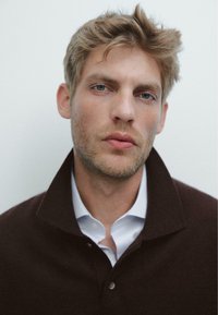 Donkerbruine gebreide polo met een hoge kraag, gedragen over een wit shirt. De achtergrond is een eenvoudige lichte kleur, die de details van het kledingstuk benadrukt.
