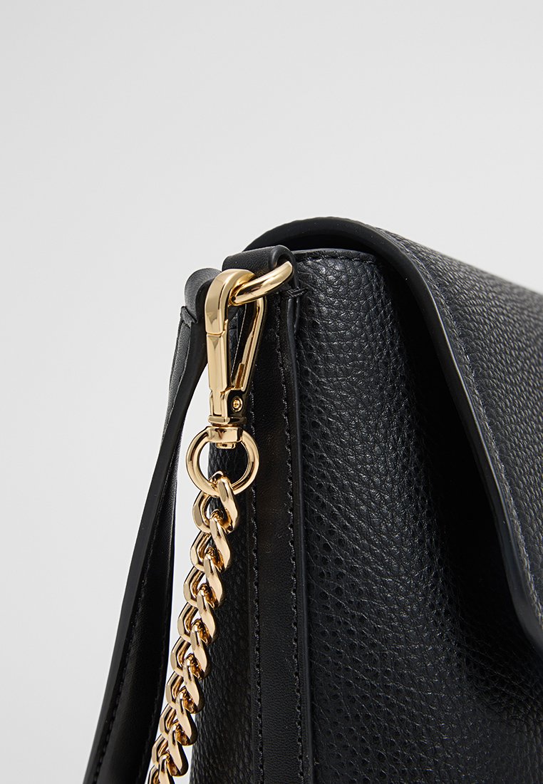 Esquina de un bolso de cuero negro texturizado con cadena dorada y detalle de cierre sobre un fondo claro.