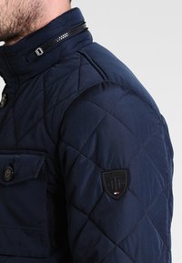 Marinblå vadderad jacka med hög krage, dragkedjestängning, bröstficka med knapp och en logotyp på vänster axel.