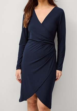 Femme portant une robe portefeuille bleu marine à manches longues, col en V et ourlet asymétrique, debout contre un mur clair uni.