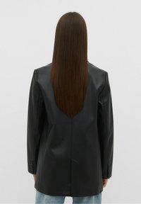 Persoon met lang, steil bruin haar in een zwarte leren blazer en lichtblauwe jeans, staand met het gezicht naar een effen lichtgrijze achtergrond.