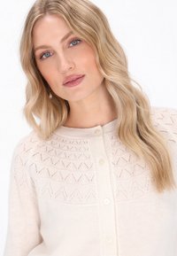 Crèmekleurige cardigan met een gestructureerd breiontwerp en geometrische patronen, ronde hals en vijf knopen aan de voorkant. Zacht, comfortabel materiaal.