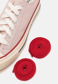 Světle růžové semišové tenisky s krémovými tkaničkami a červenými akcenty. Dvě navinuté červené tkaničky jsou položeny vedle boty.