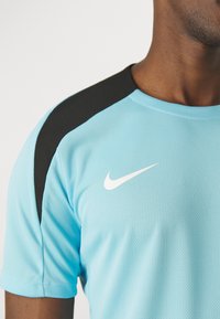 Camisa deportiva de color azul claro con acentos negros en los hombros, presentada con una textura de malla y un logotipo blanco de Nike en el pecho. Mangas cortas, cuello redondo.