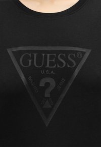 Chemise noire avec un logo triangulaire gris foncé affichant "GUESS U.S.A." et un grand point d'interrogation au centre.
