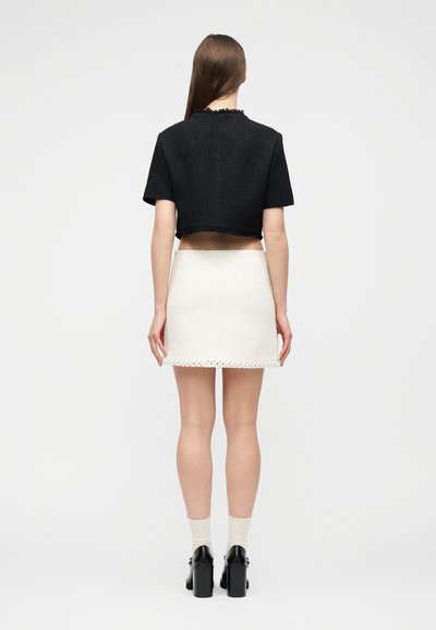 Γυναίκα με μακριά καστανά μαλλιά στέκεται γυρισμένη προς τα πίσω φορώντας μαύρο κοντό σακάκι, λευκή υφασμάτινη μίνι φούστα, λευκές κάλτσες και μαύρα παπούτσια με τακούνι.