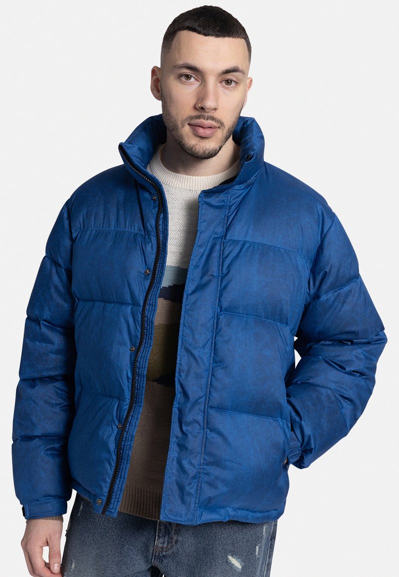 Μπλε φουσκωτό μπουφάν με ψηλό γιακά, φερμουάρ και κουμπιά κλειδώματος. Υφασμάτινος με υφή και πλισέ μοτίβο, φορεμένο πάνω από πλεκτό πουλόβερ.