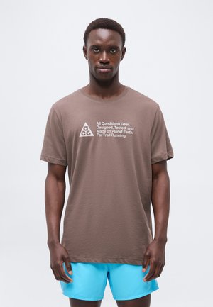 Homme portant un T-shirt marron avec le texte « All Conditions Gear » et un short bleu, debout face à un fond clair uni.