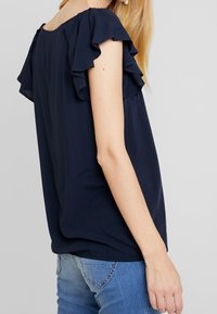 Dunkelblausa Bluse mit rüschierten kurzen Ärmeln, fließendem Design, rundem Ausschnitt und weicher Textur. Kombiniert mit Jeans.