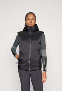 Femme portant un gilet sans manches rembourré noir sur un haut à manches longues gris avec des manches à motifs, se tenant devant un fond uni.