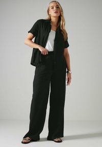 Camicia nera a maniche lunghe, ampia e con bottoni, abbinata a pantaloni larghi coordinati. T-shirt bianca sotto. Sandali piatti con motivo.