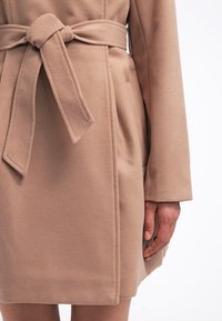 Beige uldfrakke med bundet bælte i taljen, lange ærmer og sidelommer, båret af en person med synlige hænder og ben foran en ensfarvet baggrund.