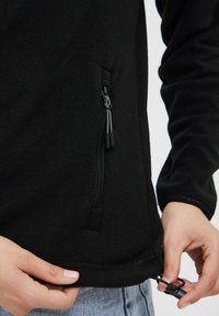 Must raehtaka featuring a zipitud tasku, reguleeritav nööriga serv ja sile tekstuur. Nähtav käsi, mis reguleerib serva.