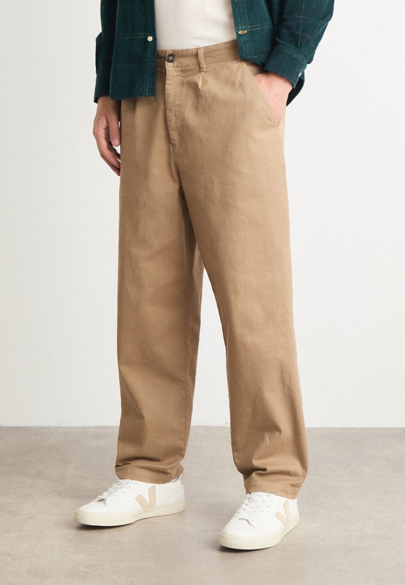 Beige Stoffhosen mit geradem Bein aus Baumwoll-Twill, ausgestattet mit Seitentaschen und einem Knopfverschluss, kombiniert mit weißen Sneakers.