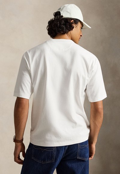 Homme portant une casquette blanche, une chemise blanche à manches courtes et un jean bleu foncé, vu de dos sur un fond neutre.