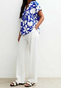 Blouse bleue à manches courtes avec un motif floral blanc, associée à un pantalon blanc à jambes larges et des sandales noires à brides blanches.