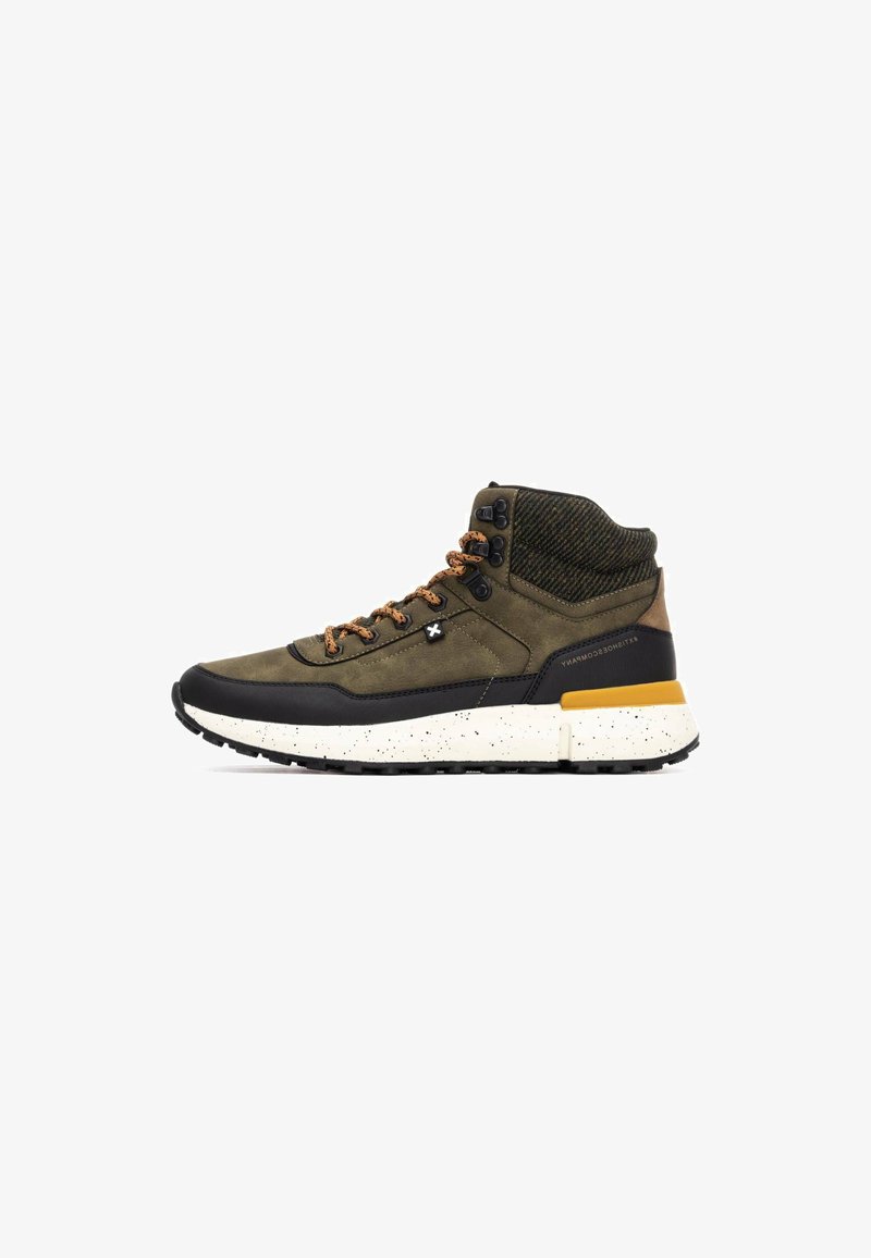 Zapatilla alta en verde oliva y negro, con tejido texturizado, cordones marrones y una suela blanca moteada con un acento amarillo.