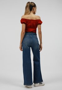 Red off-the-shoulder top with gathered sleeves paired with high-waisted blue wide-leg jeans and white sneakers with black stripes.