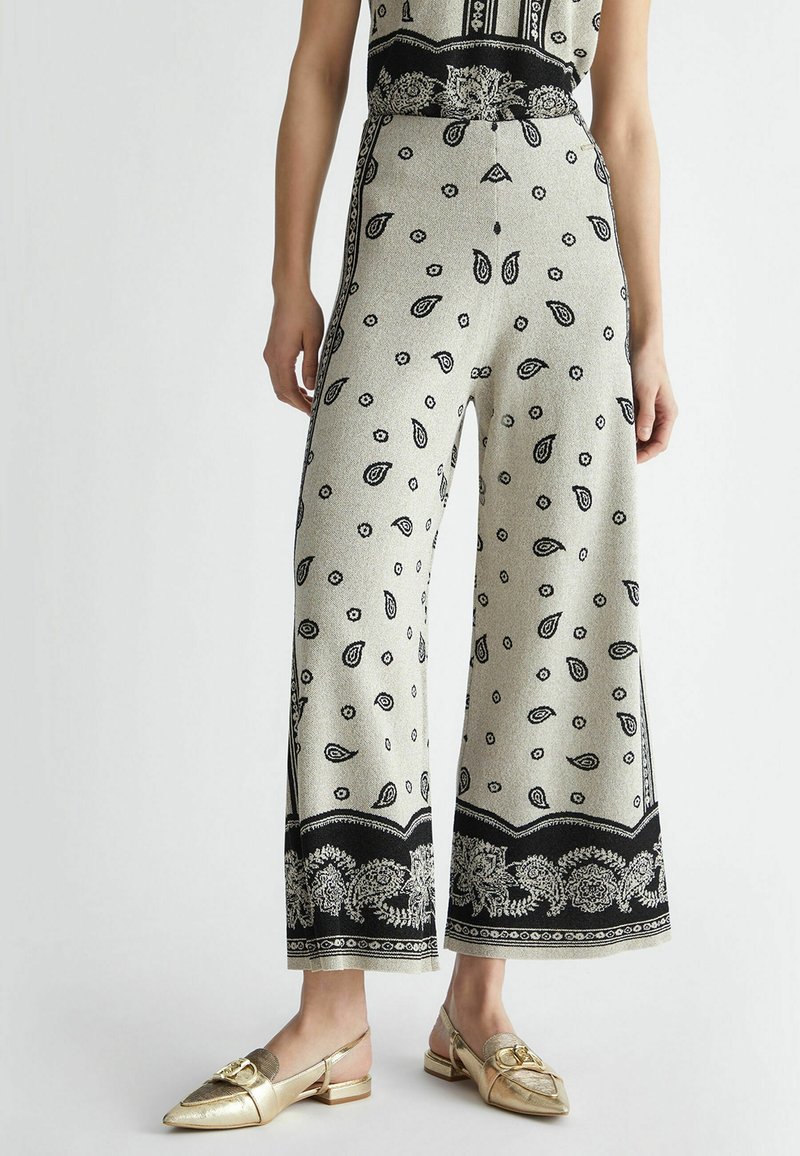Pantalon taille haute à jambes larges avec imprimé paisley noir et floral sur tissu clair, associé à des mocassins slingback dorés avec boucle métallique.