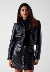 Krótka czarna skórzana kurtka z złotymi guziczkami i przednimi kieszeniami, zestawiona z pasującą czarną skórzaną mini spódnicą z zapięciem na guziki.