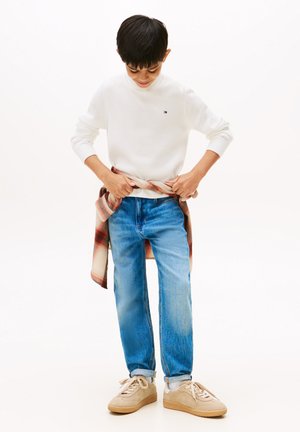 Hvit genser med rund hals og logo, lyseblå jeans og beige joggesko. Flanellskjorte knyttet rundt livet med rødt og hvitt rutemønster.