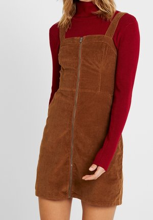 Kvinde iført brun kordfløjls-sarongkjole med lynlås foran over en rød, ribstrikket rullekravebluse med lange ærmer, hånd afslappet ned langs siden.