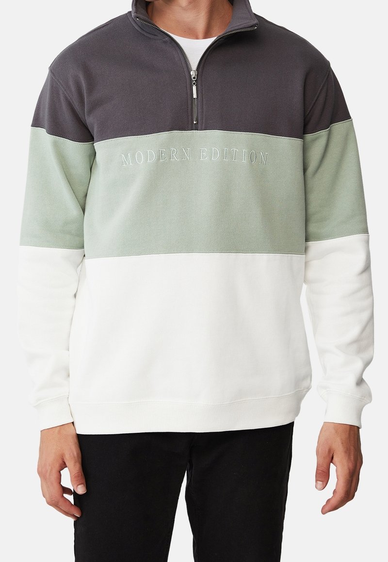Homme portant un sweat à demi-fermeture éclair trois tons avec des panneaux horizontaux noirs, vert sauge et blancs et le texte "MODERN EDITION" sur la poitrine.