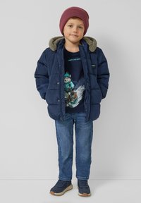 Granatowa kurtka puchowa z kapturem wyściełanym szarym futerkiem, noszona na ciemnym T-shircie z nadrukiem, w połączeniu z niebieskimi dżinsami i granatowymi trampkami.