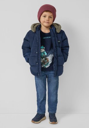 Granatowa kurtka puchowa z kapturem wyściełanym szarym futerkiem, noszona na ciemnym T-shircie z nadrukiem, w połączeniu z niebieskimi dżinsami i granatowymi trampkami.