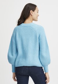 Maglione lavorato a maglia azzurro chiaro con un motivo testurizzato, maniche raglan e polsini a costine. Indossato con jeans scuri, visto da dietro.