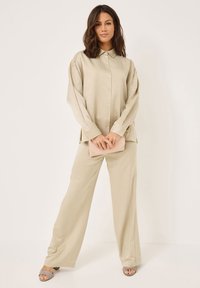 Beige, knöpfbares Hemd mit Kragen und weicher Textur, kombiniert mit locker sitzenden, beigen Hosen. Hält eine rosa Clutch. Leichtes, elegantes Design.