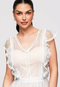 Femme aux cheveux foncés tirés en arrière, portant une robe blanche transparente à pois avec manches volantées et boucles d’oreilles créoles dorées sur fond uni.